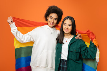 positive and trendy asian and african american women holding lgbt flag isolated on orange.
