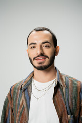 Obraz premium portrait of positive bearded man in trendy shirt and necklaces smiling at camera isolated on grey.