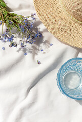White organic cotton blanket with forget me not flowers, glass of water and straw hat. Summer picnic concept composition. Copy space