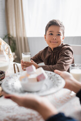 Muslim boy holding glass of milk near mother holding blurred turkish delight during ramadan in morning.