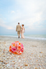 Bride and groom walking away, out of focus in background with bridal bouquet in the foreground on sandy beach