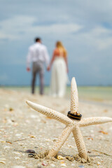 Wedding rings on star fish with blurred wedding couple walking away in background on the beach.