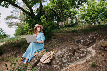 A young girl freelancer works with a laptop in nature. Dressed with a blue summer dress and a light hat. Sits on stones in a forested area. Nearby lies a beige leather bag.