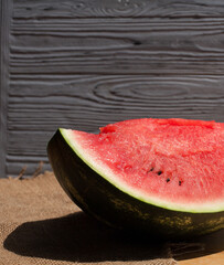 A ripe large piece of a red cut striped watermelon with pits lies on a cloth, on a light yellow wooden table, against the background of a blue textured wooden wall.