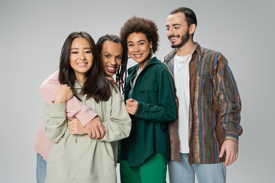 Trendy Multiracial Man With Dreadlocks Embracing Asian Woman And Smiling At Camera Near Happy Friends Isolated On Grey.