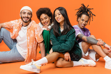 happy multiethnic friends in trendy attire sitting and smiling at camera on orange background.