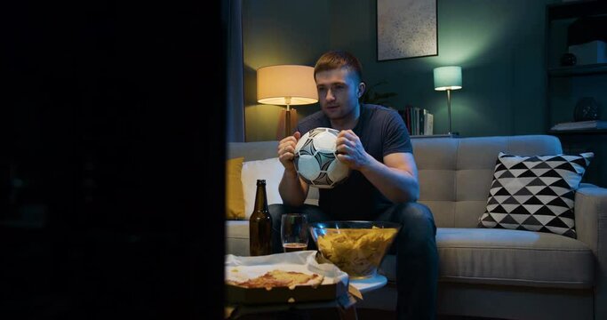 Caucasian male football fan watching sport match on TV alone at home late at night. Dissapointed guy worrying because his favorite team missing goal or loosing match. Man watching sport channel.