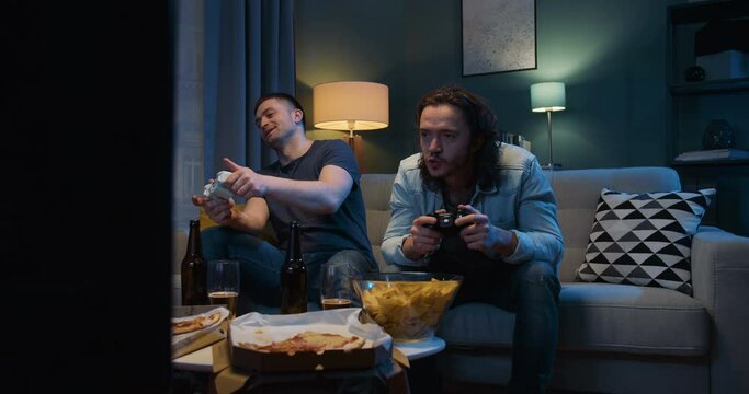 Two Caucasian friendly guys playing videogame with joystick while sitting in tension late in evening with snacks and beer in front of TV. Men having fun together with games.