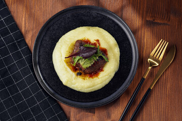 A medium rare ostrich steak in barbecue sauce on a cloud of herb mashed potato topped with fresh green arugula leaves on a black plate on a wooden background, top view