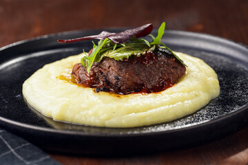 A medium rare ostrich steak in barbecue sauce on a cloud of herb mashed potato topped with fresh green arugula leaves on a black plate on a wooden background