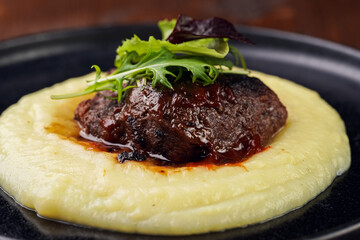 A medium rare ostrich steak in barbecue sauce on a cloud of herb mashed potato topped with fresh green arugula leaves on a black plate on a wooden background