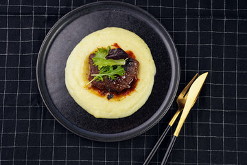 A medium rare ostrich steak in barbecue sauce on a cloud of herb mashed potato topped with fresh green arugula leaves on a black plate on a black checkered table cloth, top view