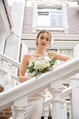 low angle view of bride in white dress holding bouquet and looking at camera near house.