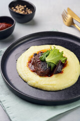 A medium rare ostrich steak in barbecue sauce on a cloud of herb mashed potato topped with fresh green arugula leaves on a black plate on a grey background
