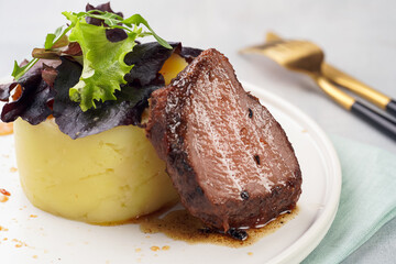A medium rare ostrich steak in barbecue sauce with herb mashed potatoes topped with fresh green arugula leaves on a white plate on a grey background