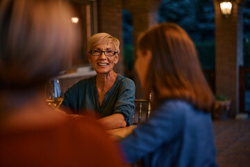 Happy senior woman talks to her daughter and granddaughter during family dinner on patio.