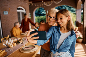 Happy teenage girl and her grandmother taking selfie during family gathering on patio.