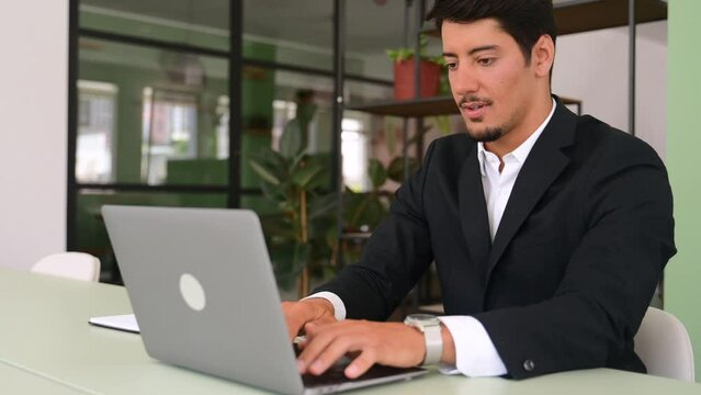 Excited lucky hispanic young businessman sitting at the desk in front of laptop in office and rejoicing with good news, overjoyed male office employee celebrating job offer, clenched fists in triumph
