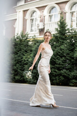 Fototapeta premium full length of young bride in white dress holding wedding bouquet of blooming flowers and walking outside.