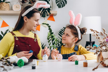 Happy young mother with smiling little kid daughter to dye and decorate eggs with paints for Easter holidays while sitting together at white table in room. Easter Family traditions and spring concept