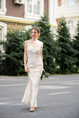 full length of pretty bride in white dress holding wedding bouquet and walking on street.