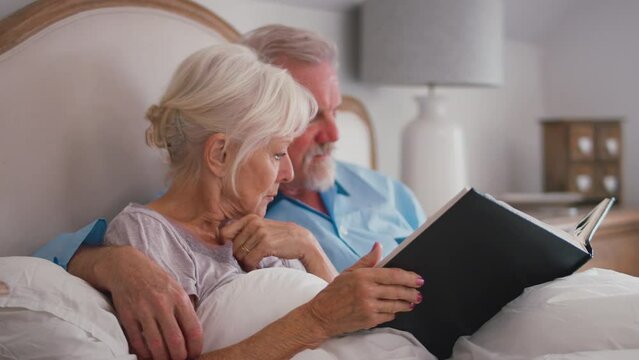 Smiling retired senior couple in bed at home looking through photo album and reliving old memories - shot in slow motion