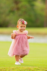 1 year old baby girl in a pink dress, walking on green grass.