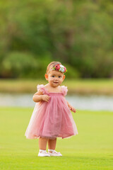1 year old baby girl in a pink dress walking on green grass.