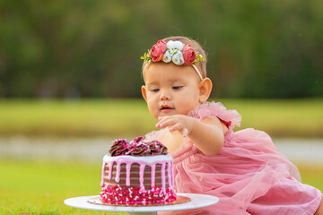 1 year old baby girl sitting behind birthday cake outdoors on green grass.