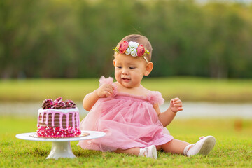 1 year old baby girl sitting behind birthday cake outdoors on green grass.