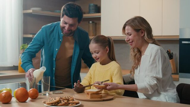 Caucasian family cooking breakfast together at kitchen at home mother with little daughter child girl prepare toasts bread with chocolate pasta father pour milk into glasses parents with kid cook