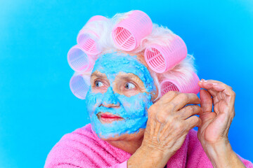 woman with blue cream moistening mask on face , puts curlers on white hair and wear pink bath robe in studio background