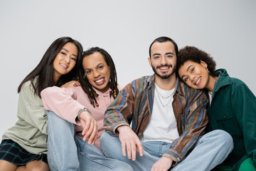 cheerful multicultural friends in trendy outfit smiling at camera while sitting isolated on grey.