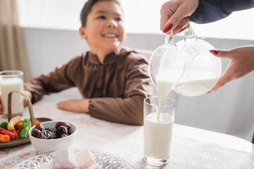 Muslim mother pouring milk during suhur breakfast near blurred son at home.
