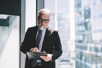Senior businessman standing with clipboard near glass wall