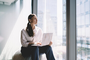 Thoughtful woman sitting with laptop near glass wall