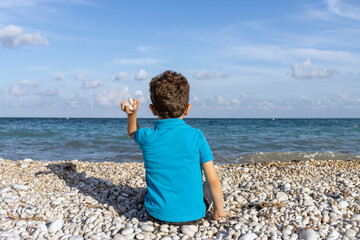 child sitting on the shore of the beach throwing stones into the water seen from behind