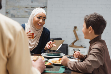Fototapeta na wymiar African american woman in hijab holding tea and talking to son near husband and food at home.