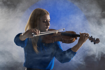 Beautiful blond girl playing violin. Female violinist against dark blue background with smoke.