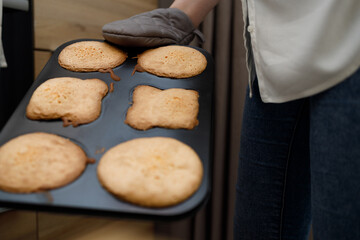Woman holding hot tray with baked cupcakes, muffins, cookies in oven-glove near oven. No face