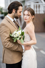 bearded groom hugging bride in wedding dress holding blooming flowers.