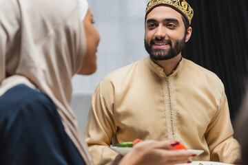 Positive muslim man looking at blurred wife with food at home.