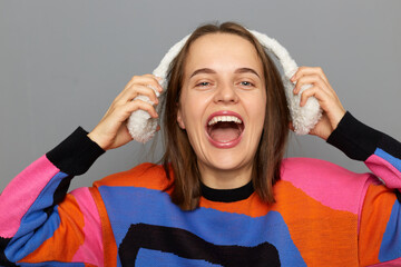 Portrait of smiling cute pretty happy woman wearing colorful jumper and earmuffs, standing isolated on gray background, looking at camera and screaming with happiness.