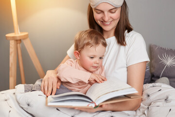 Indoor shot of smiling european woman wearing white t-shirt and blindfold sitting in bed with her toddler daughter, mommy reading fairy tale for kid, holding book in hands.