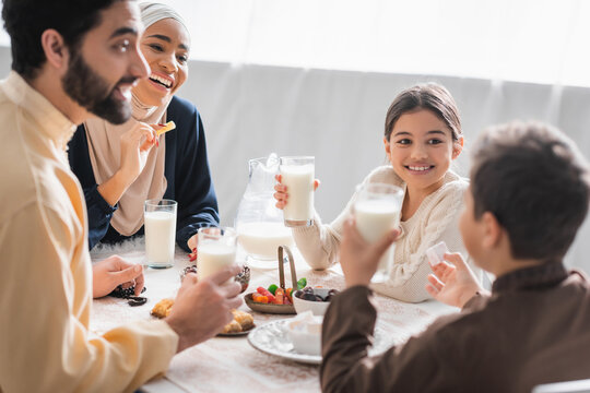 Positive muslim family holding glasses of milk and talking near food during suhur at home.