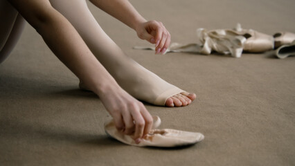 Beautiful ballet dancer sits on floor near pointe at dance studio and puts on ballet shoes before choreography lesson. Ballerina prepares for performance. Classical ballet dance school. Feet close up.