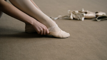 Ballerina sits on floor at dance studio and puts on pointe shoes before choreography lesson. Graceful ballet dancer prepares for performance or training. Classical ballet dance school. Feet close up.