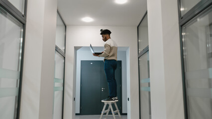 African american installer sets up security cameras in office room using laptop. Man stays on...