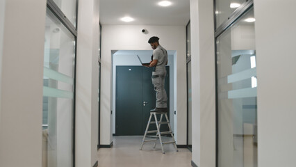 African american installer sets up security cameras in office room using laptop. Man stays on...