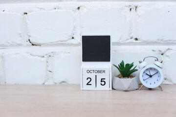 Wooden calendar with date October 25 and alarm clock with plant on table against brick wall...